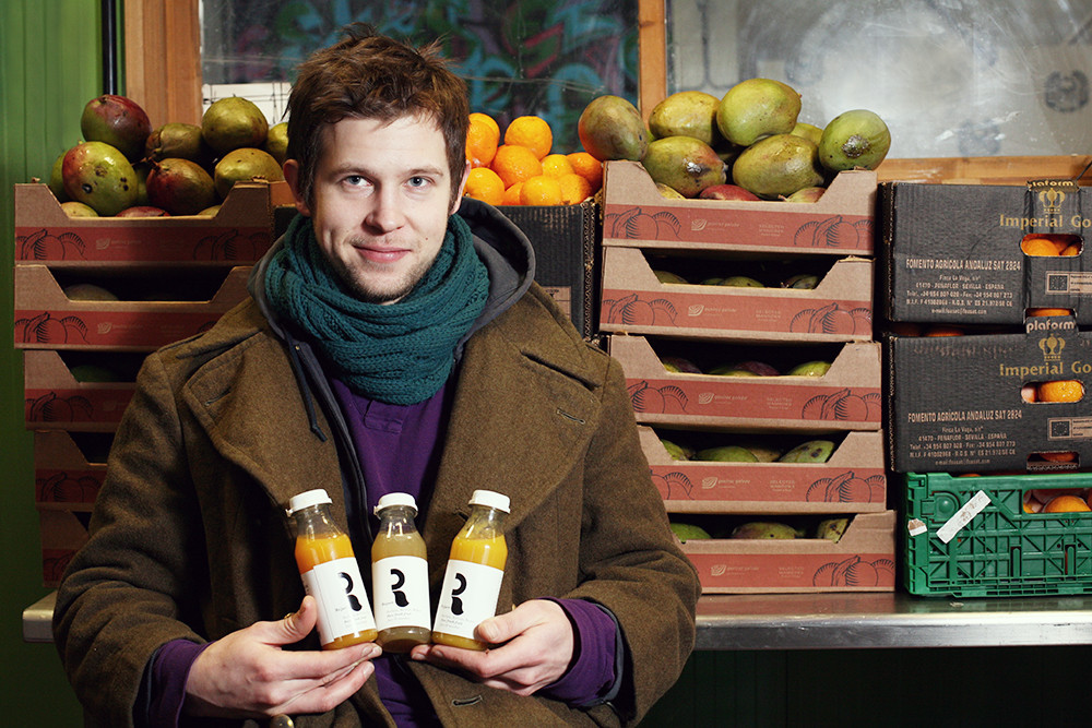 Man wearing a scarf stands in front of wooden food bin holding three bottles of fresh made juices